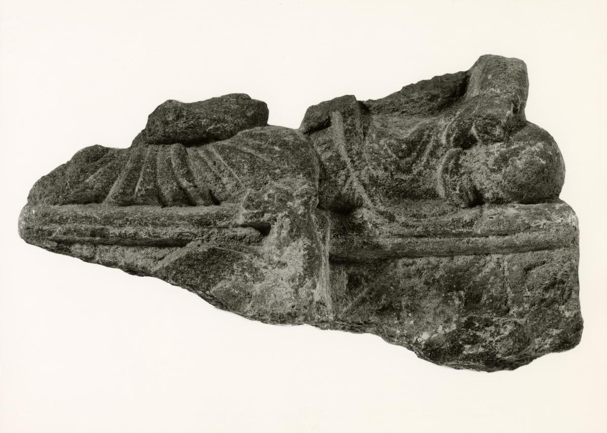 Erzählende Szene (Fragment): Unterkörper eines sitzenden Buddha (?) mit aufbäumendem Tier (?) im Sockel (links, erste Predigt?); sitzender Mönch (?) (rechts)