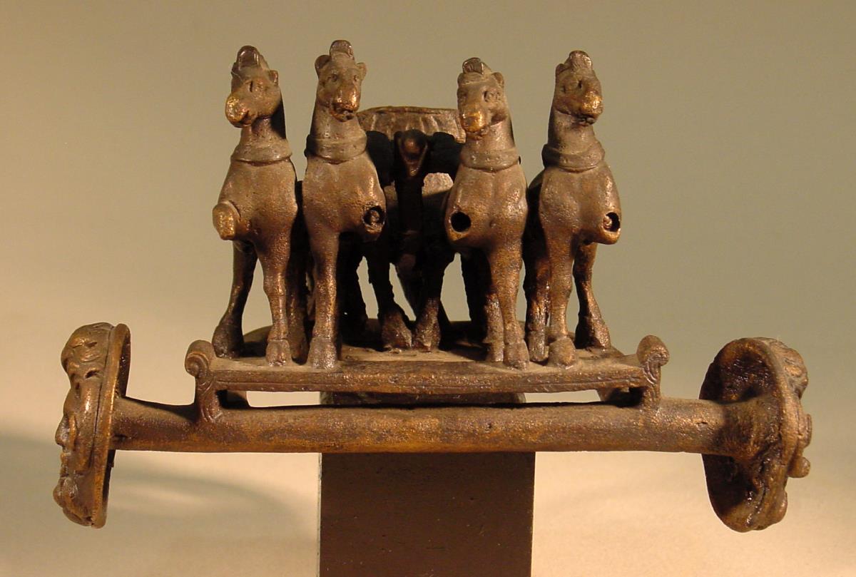 Gerätteil in Form einer Quadriga, reduziert aber gut erhalten, verloren sind die vier vorgestreckten Beine der Pferde, anstelle der Räder Scheiben mit Ammonsköpfen