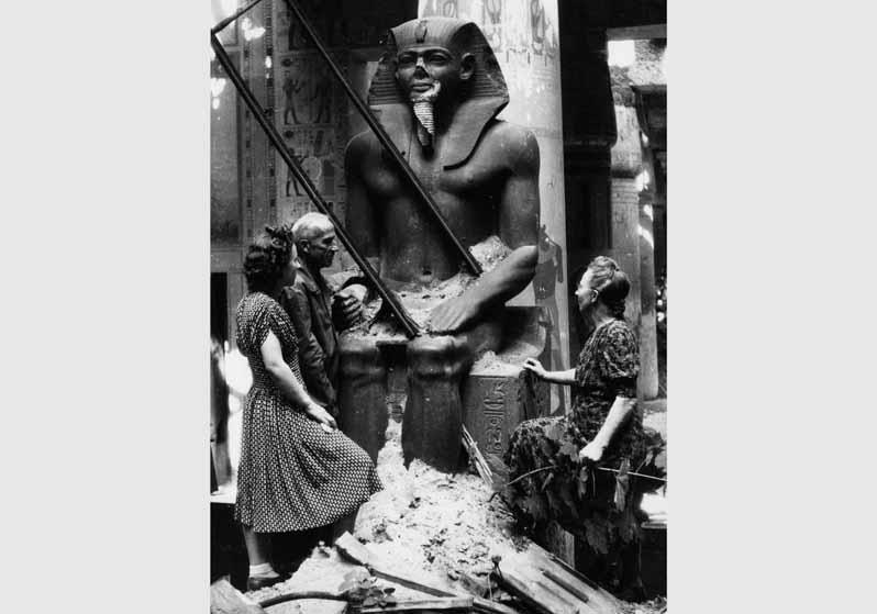Restaurator Rudolf Kuhn mit zwei Frauen an der Kolossalstatue Sesostris II im Ägyptischen Hof im Neuen Museum