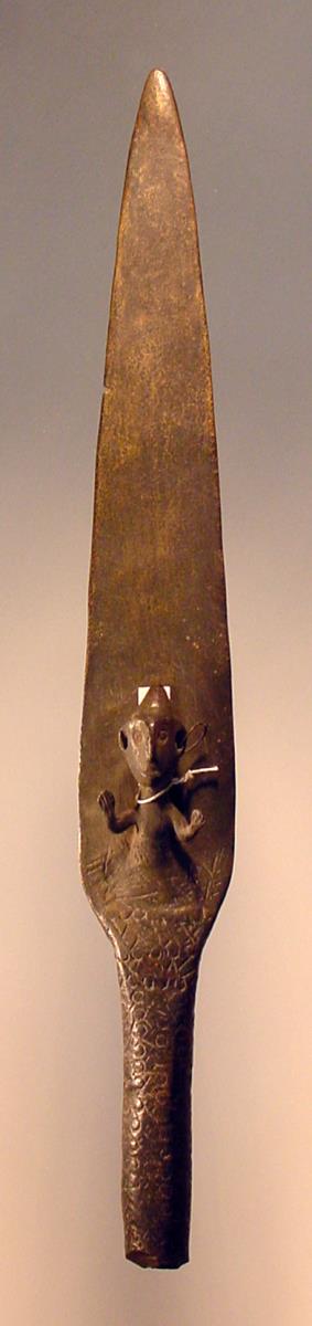Große phönizische Lanzenspitze mit langem massivem Fortsatz. Röhre abgehauen. Unten mit halber menschlicher Figur mit konischer Mütze, Hände nach oben