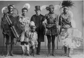 Gruppe von vier bewaffneten Zulukriegern und ein Kind sowie ein Herr mit Frack und Zylinder (OT)