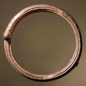 Massiver runder Armring von rundem Querschnitt mit übergreifenden Enden; an der Außenseite mit sechs kannelurenartigen Rillen; an den Enden mit runden Knöpfen versehen, vor welchen halbumlaufende Einkerbungen