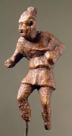 Römische Statuette eines Sklaven oder Possenreiβers (?). Kurzer Chiton, der von der linken Schulter niedergleitet. Rechte Hand mit sprechendem Gestus vorgestreckt. Kurzgeschnittene Haare mit Schopf. Beide Füße weggebrochen, r. Hand beschädigt. Rauhe Patina