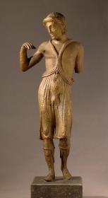 Klassische Statuette eines Jünglings mit einem den Unterkörper umkleidenden Chiton, welcher durch zwei Stricke über den Schultern gehalten wird.